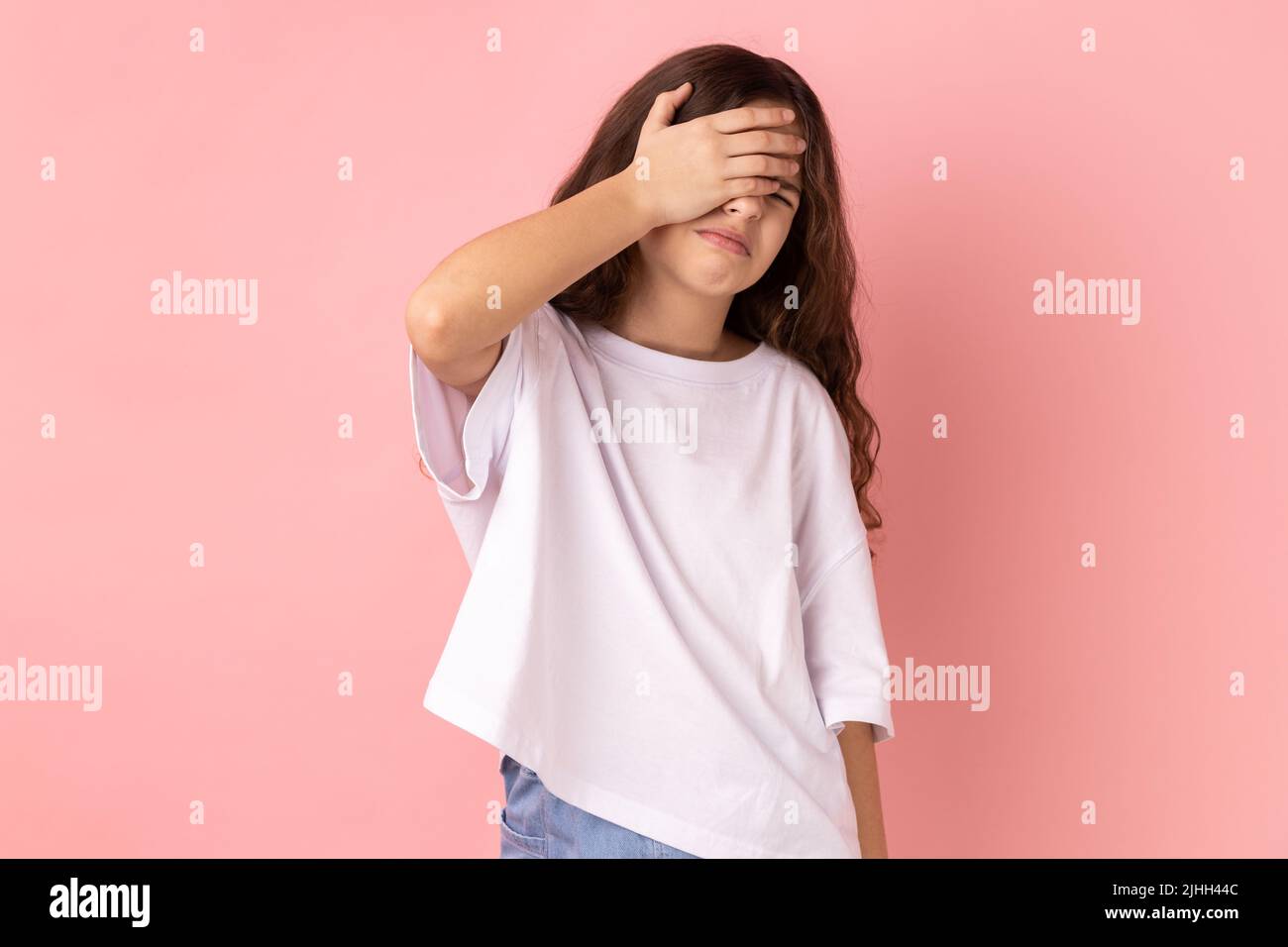 Non vuoi guardare questo. Ritratto di bambina che indossa una T-shirt bianca che copre gli occhi con la mano, sentendosi shamed e spaventato guardare. Studio interno girato isolato su sfondo rosa. Foto Stock