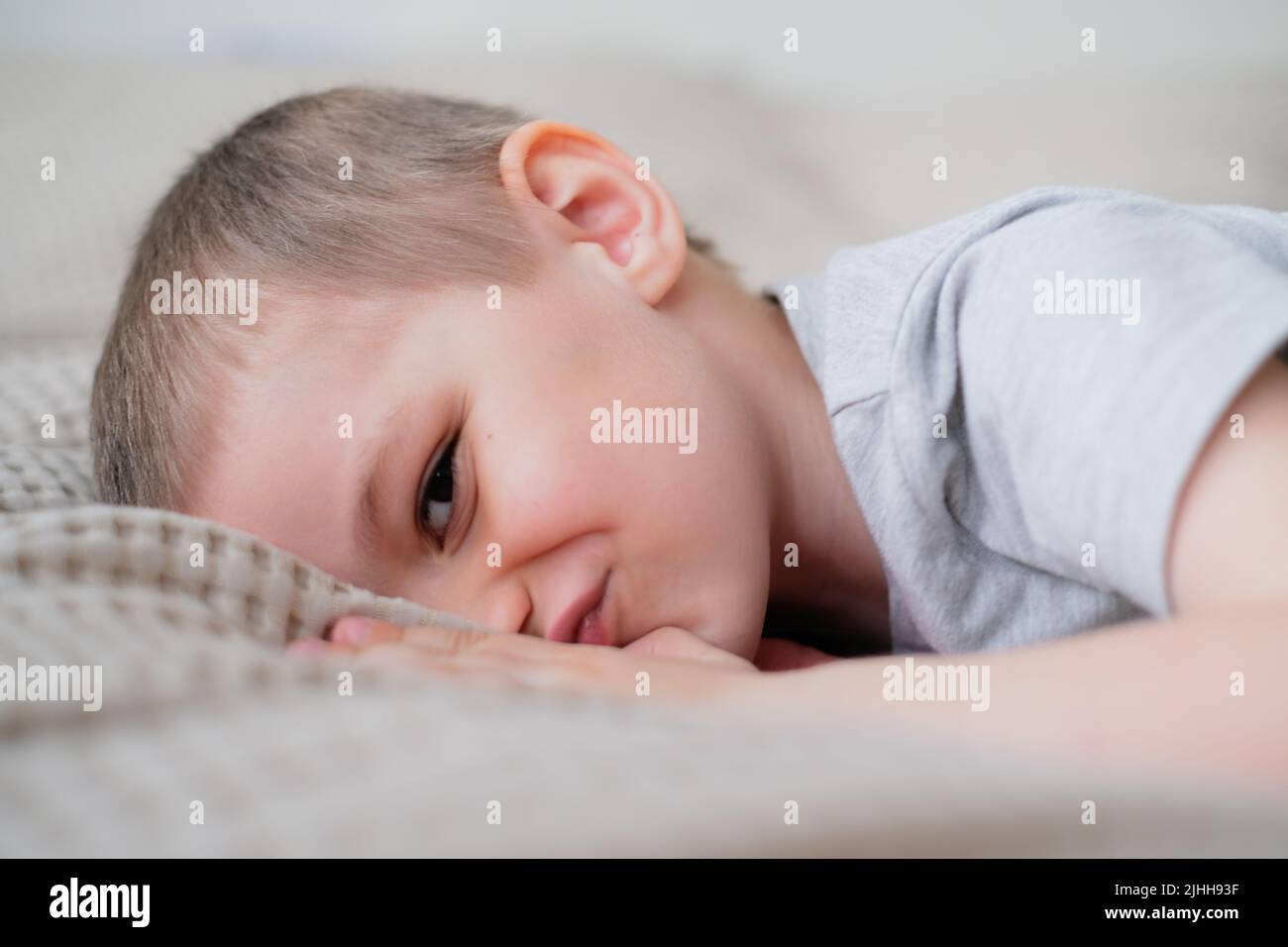 Primo piano di un piccolo ragazzo caucasico felice in una t-shirt grigia si trova sul letto e sorride Foto Stock