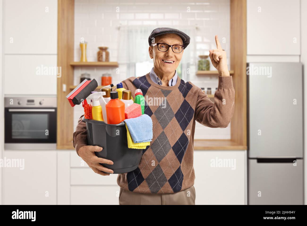 Uomo anziano che tiene un secchio con i rifornimenti di pulizia e che punta in su all'interno di una cucina Foto Stock