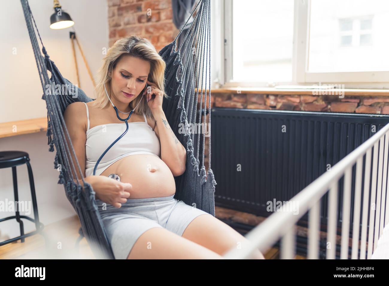 Donna caucasica incinta con capelli biondi colorati e grande ventre di gravidanza seduta su un'oscillazione interna e toccando il suo ventre con uno stetoscopio per sentire il battito del suo feto. Foto di alta qualità Foto Stock