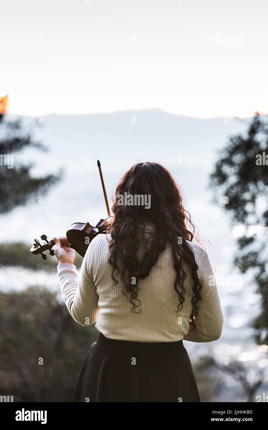 Violinista femminile di bruna visto dalla parte posteriore che suona il violino fuori nella montagna. Verticale Foto Stock