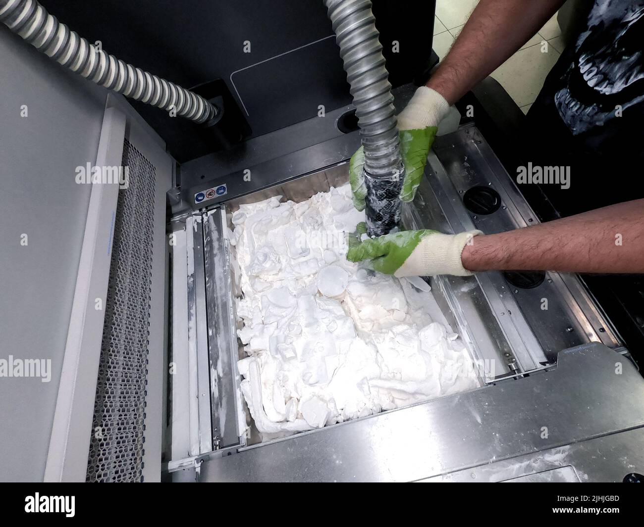 Un uomo che lavora con un aspirapolvere funzionante per pulire la polvere bianca di poliammide da un modello stampato su una stampante 3D all'interno di una stampante 3D. Pulizia di oggetti stampati su una stampante industriale a polvere 3D Foto Stock