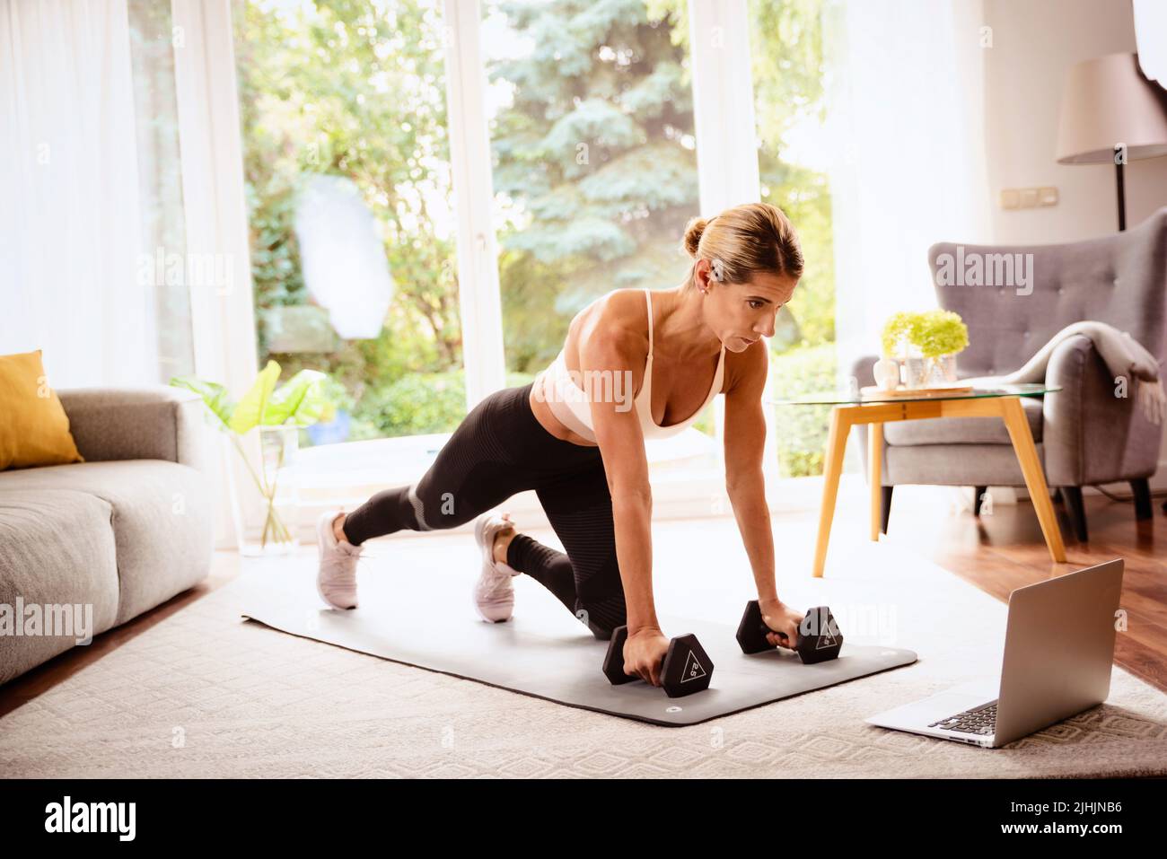 Colpo di una donna usando i dumbbells mentre fa l'allenamento del corpo a casa. Donna che indossa abbigliamento sportivo e utilizza un computer portatile mentre si esercita sul tappetino yoga. Healt Foto Stock