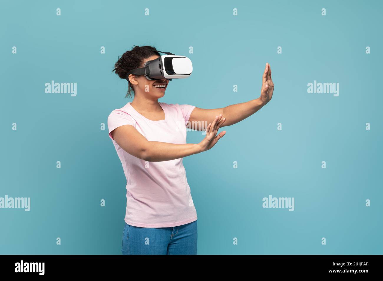Gioiosa donna nera nel visore VR che tocca l'aria durante l'esperienza di realtà virtuale su sfondo blu, spazio libero Foto Stock