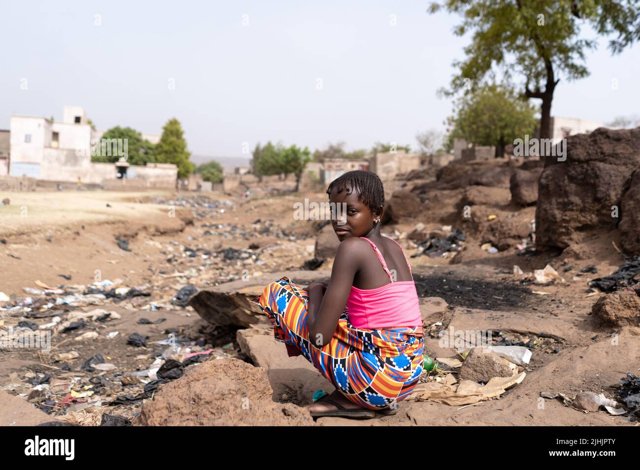 Giovane ragazza africana seduta sulla riva di un letto di fiume eroso drenato pieno di rifiuti e sporcizia; concetto di riscaldamento globale e inquinamento Foto Stock