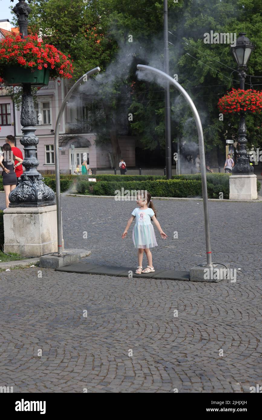 Le ragazze piccole carine amano il raffreddamento dai nebulizzatori pubblici su una piazza della città; raffreddamento della nebbia dell'acqua, spruzzatori d'acqua Foto Stock