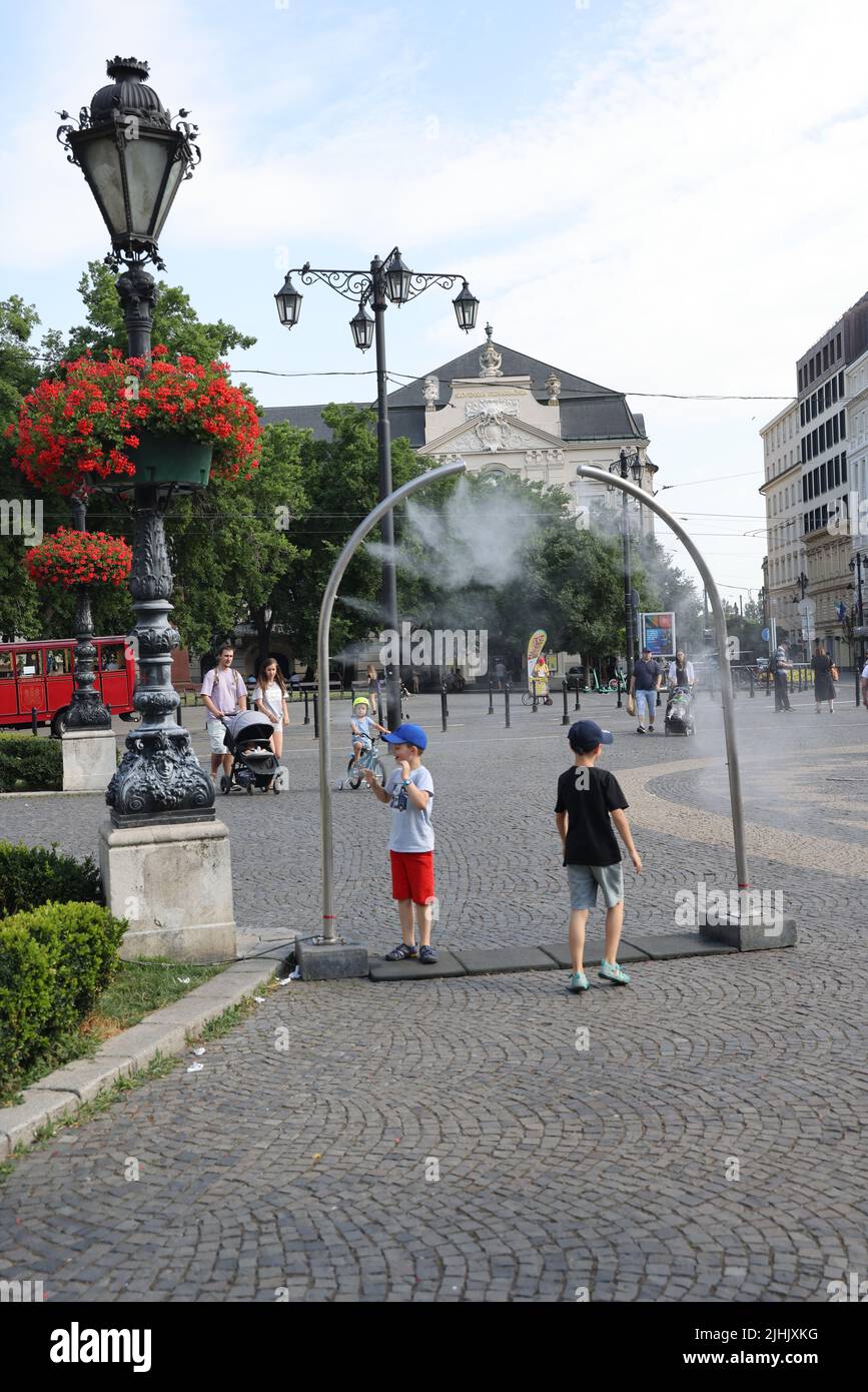 Due bambini si godono rinfreschi da nebulizzatori pubblici su una piazza della città; acqua nebulizzata raffreddamento, nebbia raffreddamento, acqua spruzzatori Foto Stock