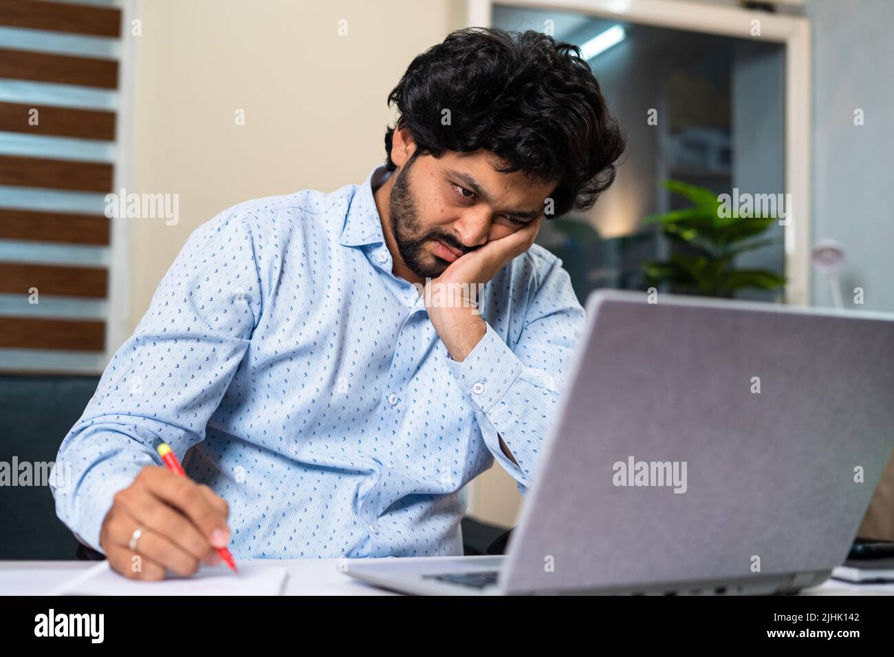 Giovane uomo stanco mentre si lavora su un notebook prendendo appunti alla scrivania dell'ufficio - concetto di frustrazione del lavoro, vita lavorativa di routine e esaurimento. Foto Stock