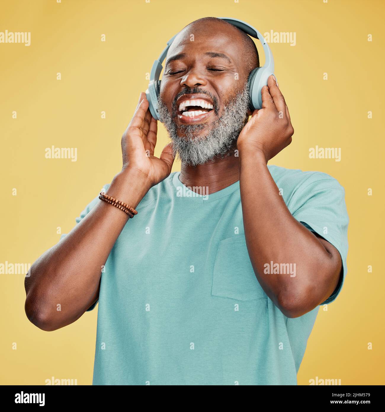 Un uomo afro-americano maturo e felice in piedi da solo su uno sfondo giallo in uno studio e indossando le cuffie per ascoltare la musica. Ritratto sorridente Foto Stock