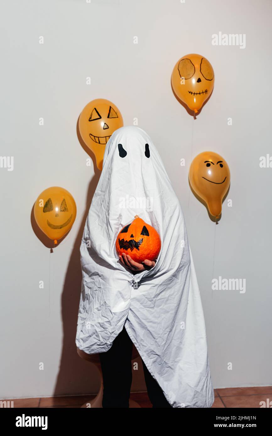 Buon Halloween. Ghost tiene in mano la zucca arancione. Bambino in abito bianco, palloncini gialli dipinti. Emozioni diverse di gioia, rabbia, risate. Design festivo, concetto di festa. Messa a fuoco selettiva morbida Foto Stock