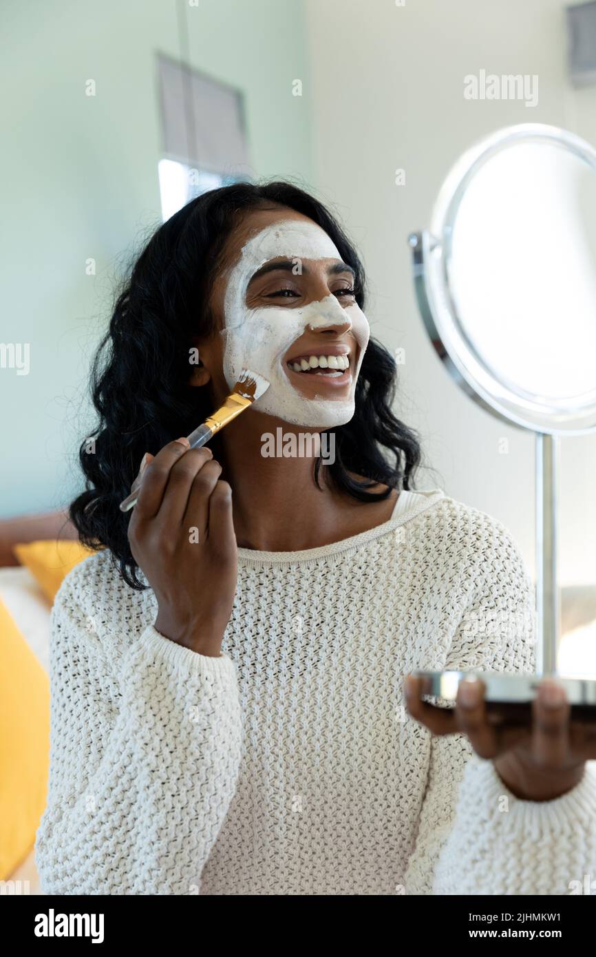 Sorridendo giovane donna biraciale applicare crema di bellezza con pennello sul viso mentre si guarda nello specchio Foto Stock