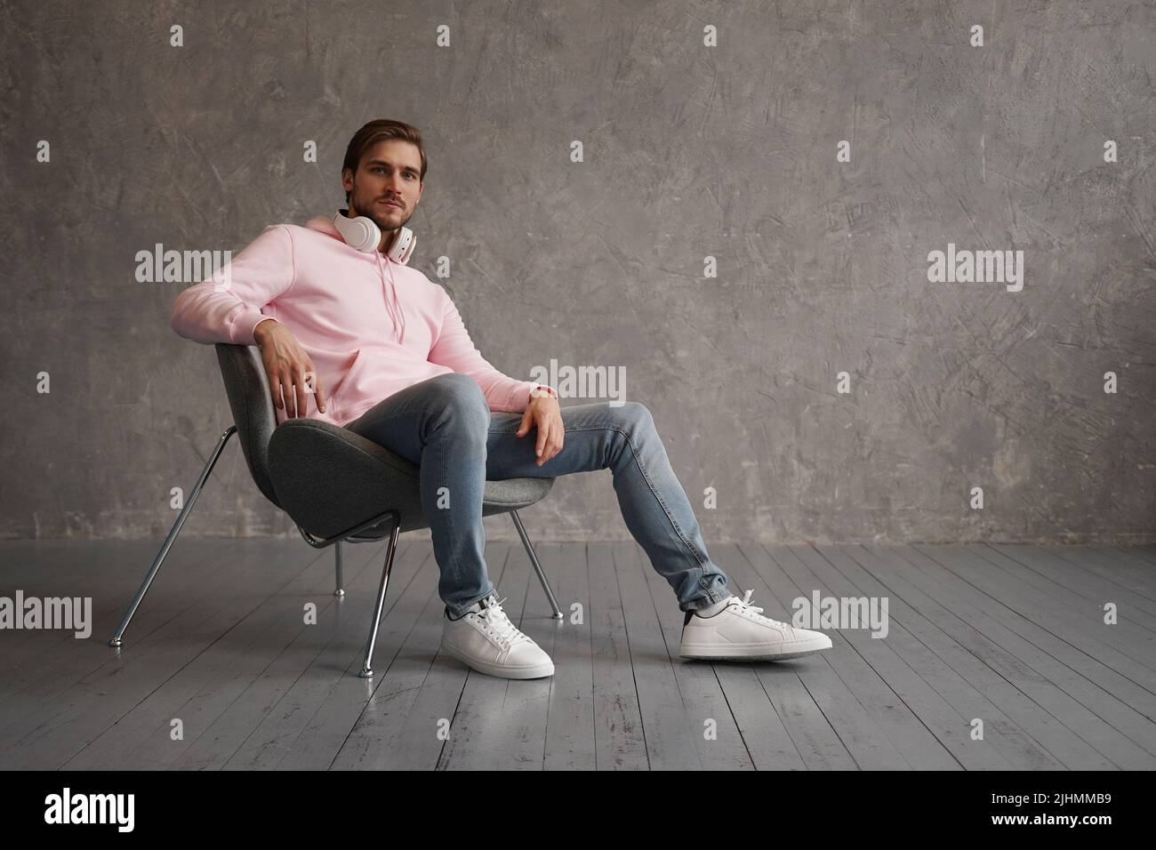 Sentirsi a proprio agio ovunque. Tutta la lunghezza del bel giovane uomo che guarda la macchina fotografica mentre si siede su sfondo grigio Foto Stock