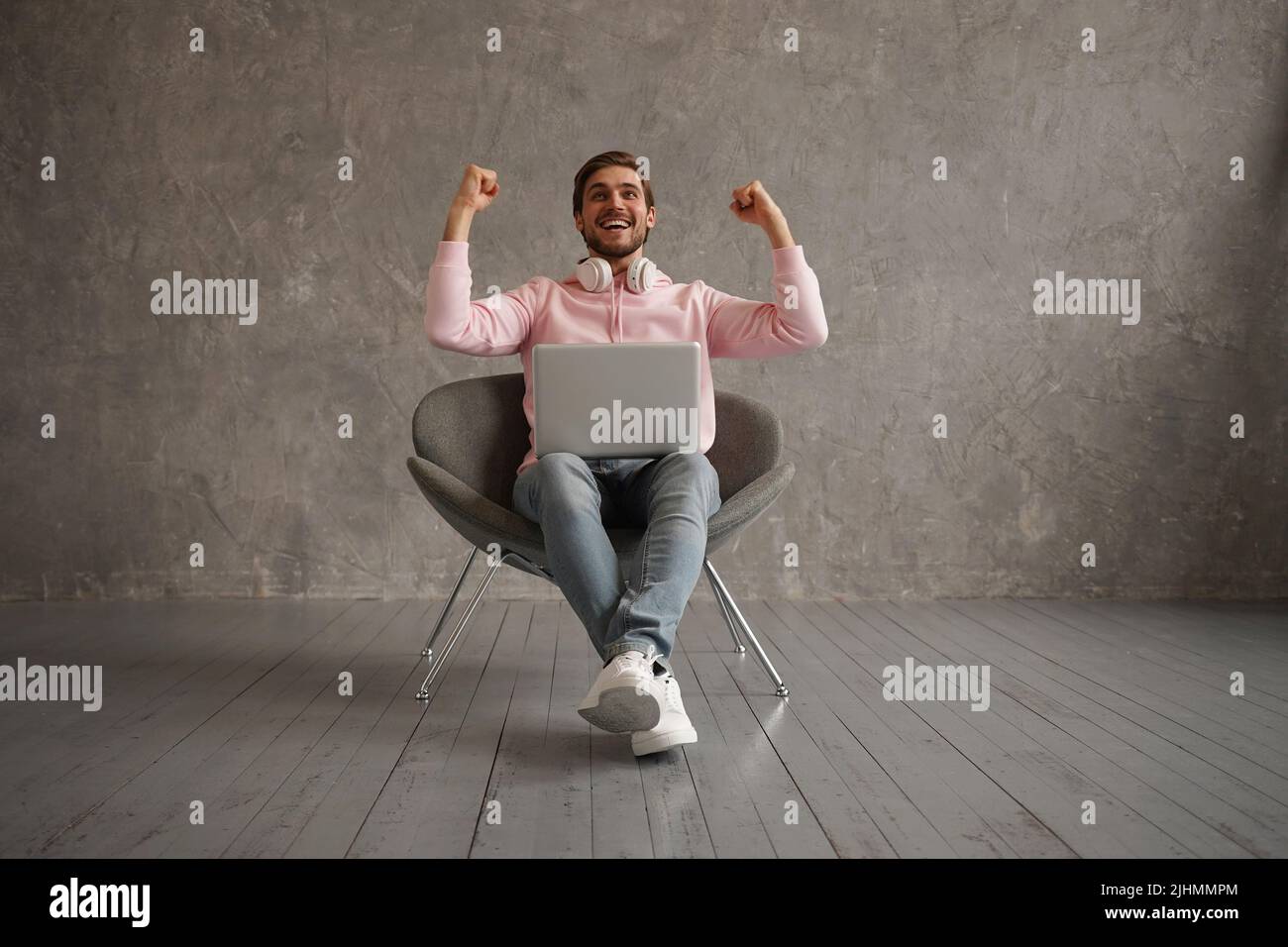 Maschio gioioso usando il laptop e scuotendo i Fists che festeggiano le grandi notizie e la fortuna seduta nella sedia sopra sfondo grigio della parete Foto Stock