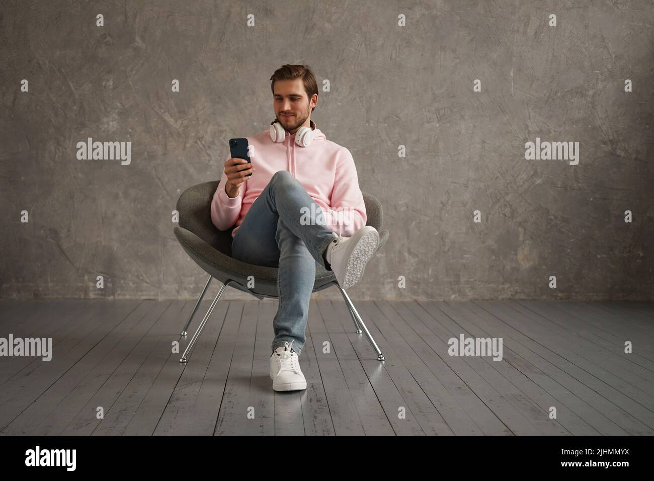 Il ragazzo allegro che usa il telefono mobile con la nuova applicazione seduta nella poltrona sopra sfondo grigio della parete Foto Stock