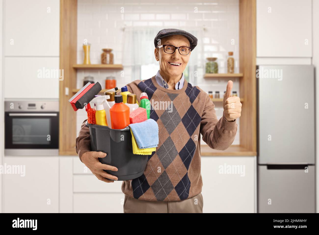 Uomo anziano che tiene un secchio con i rifornimenti di pulizia e che gestura i pollici in su all'interno di una cucina Foto Stock