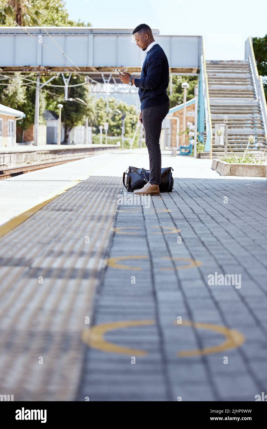 Uomo d'affari nero che viaggia da solo. Un giovane uomo d'affari afroamericano in attesa di un treno in una stazione ferroviaria e utilizzando il suo cellulare wireless Foto Stock