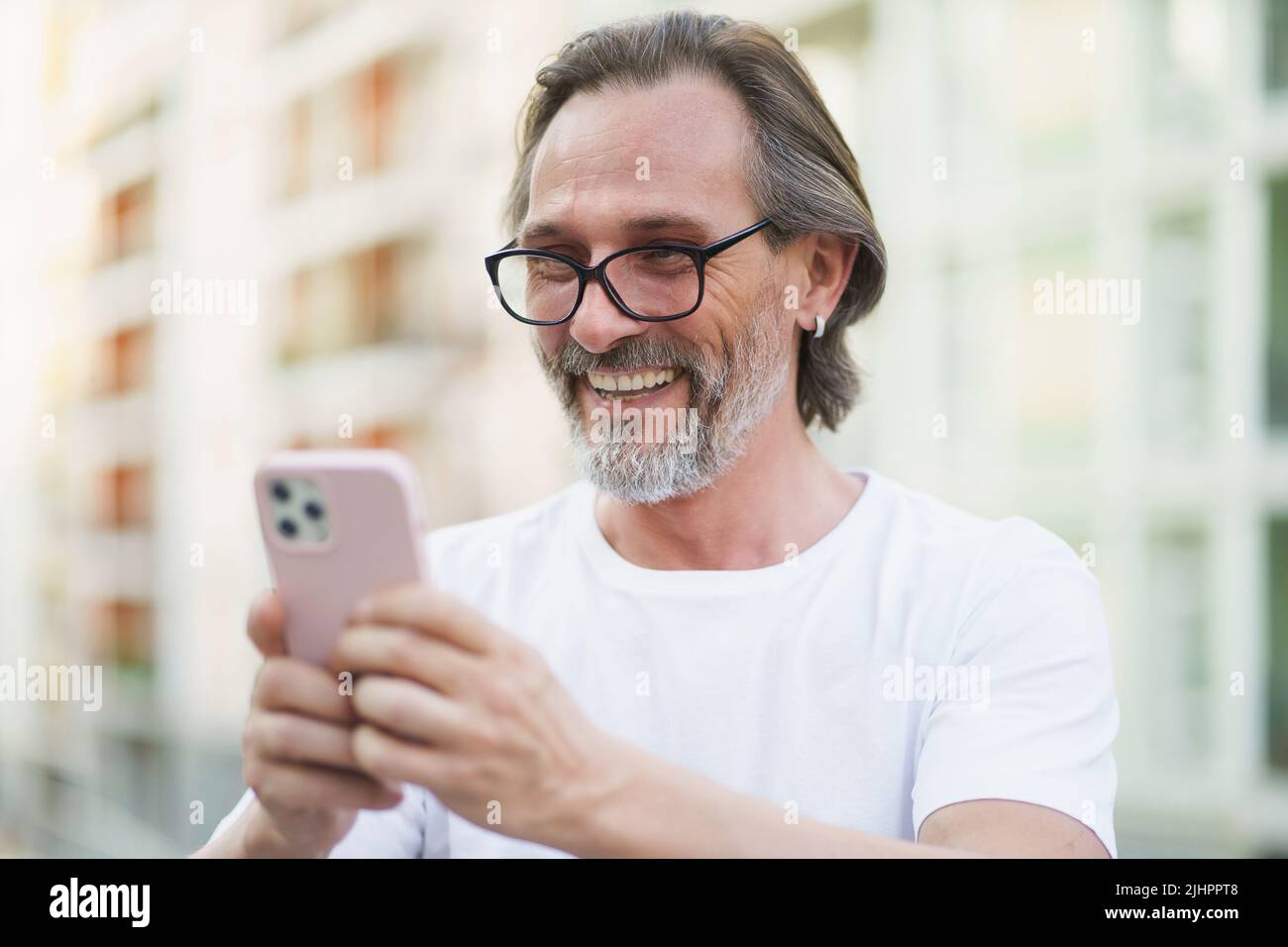 Felice uomo caucasico di mezza età con barba grigia lettura o SMS utilizzando smartphone in piedi all'aperto strade della città sfondo indossando camicia bianca. Uomo di mezza età per strada della città europea. Foto Stock