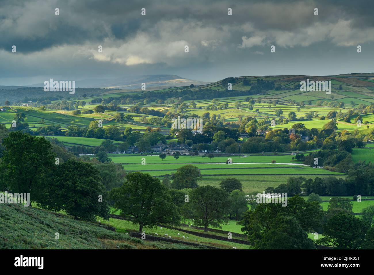 Panoramica Burnsall in valle (pendii collinari, altipiani, campi verdi e pascoli, pareti di pietra secca, cielo nuvoloso e sole) - Upper Wharfedale, Inghilterra, Regno Unito. Foto Stock