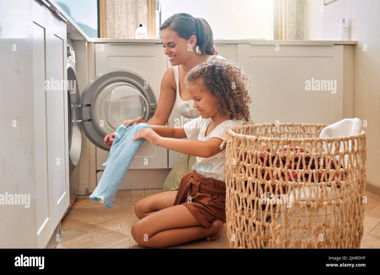 La giovane madre ispanica e sua figlia che smistano la biancheria sporca nella lavatrice a casa. Adorabile bambina e sua madre che fanno i compiti insieme Foto Stock