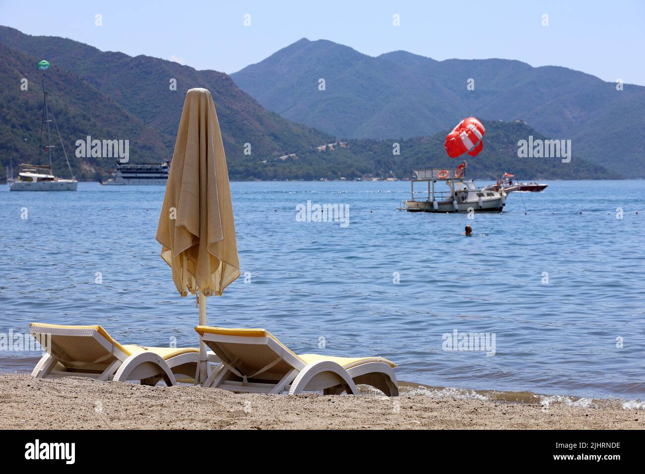 Sedie a sdraio e ombrellone vuoti su una spiaggia. Vista sul Mar Mediterraneo, barca con parasailing e verdi montagne in nebbia Foto Stock