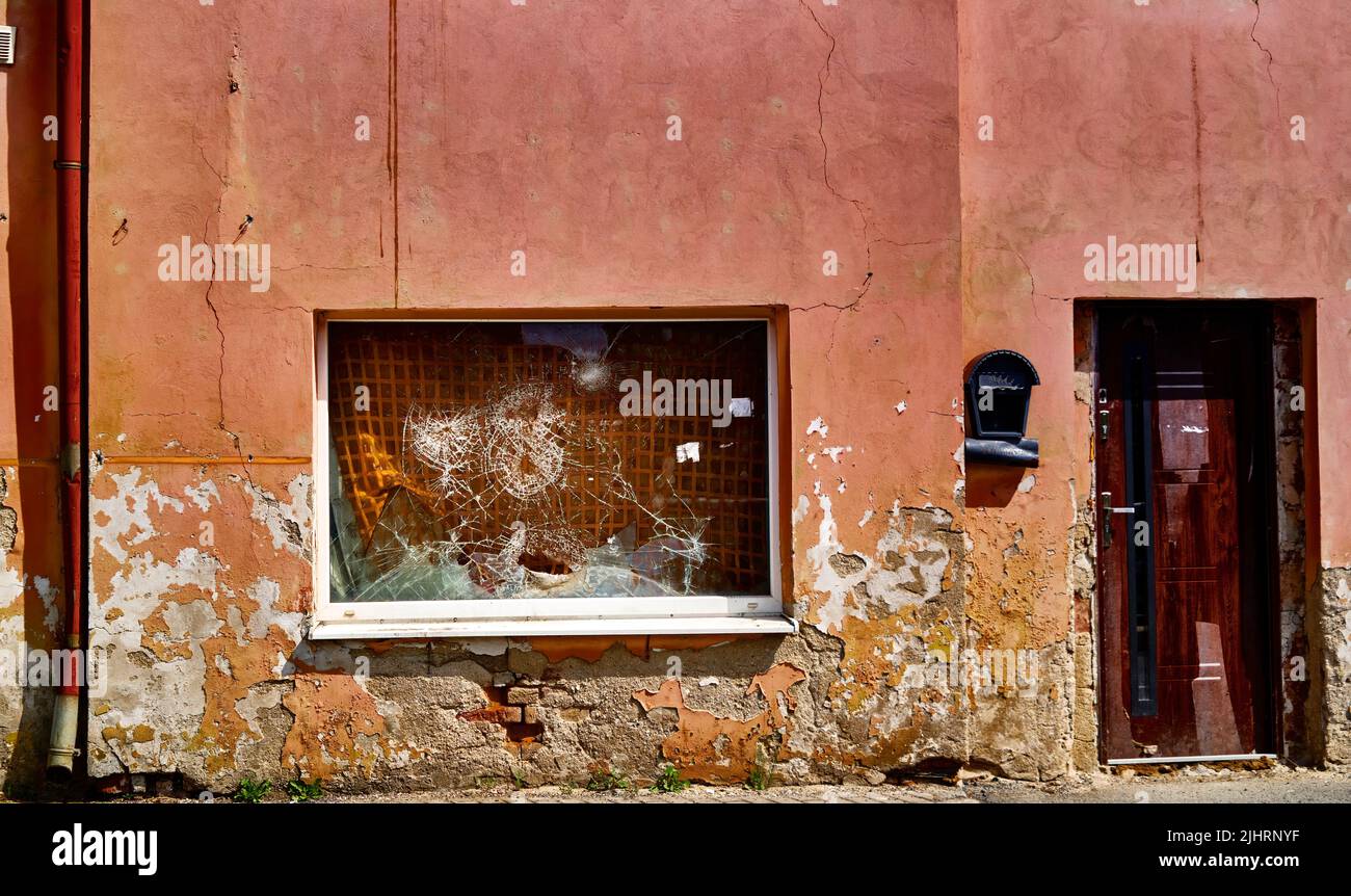 Sbucciare la vernice su una facciata dilapidata con un gocciolatoio, una porta di legno e una vetrina il cui vetro è rotto più volte Foto Stock