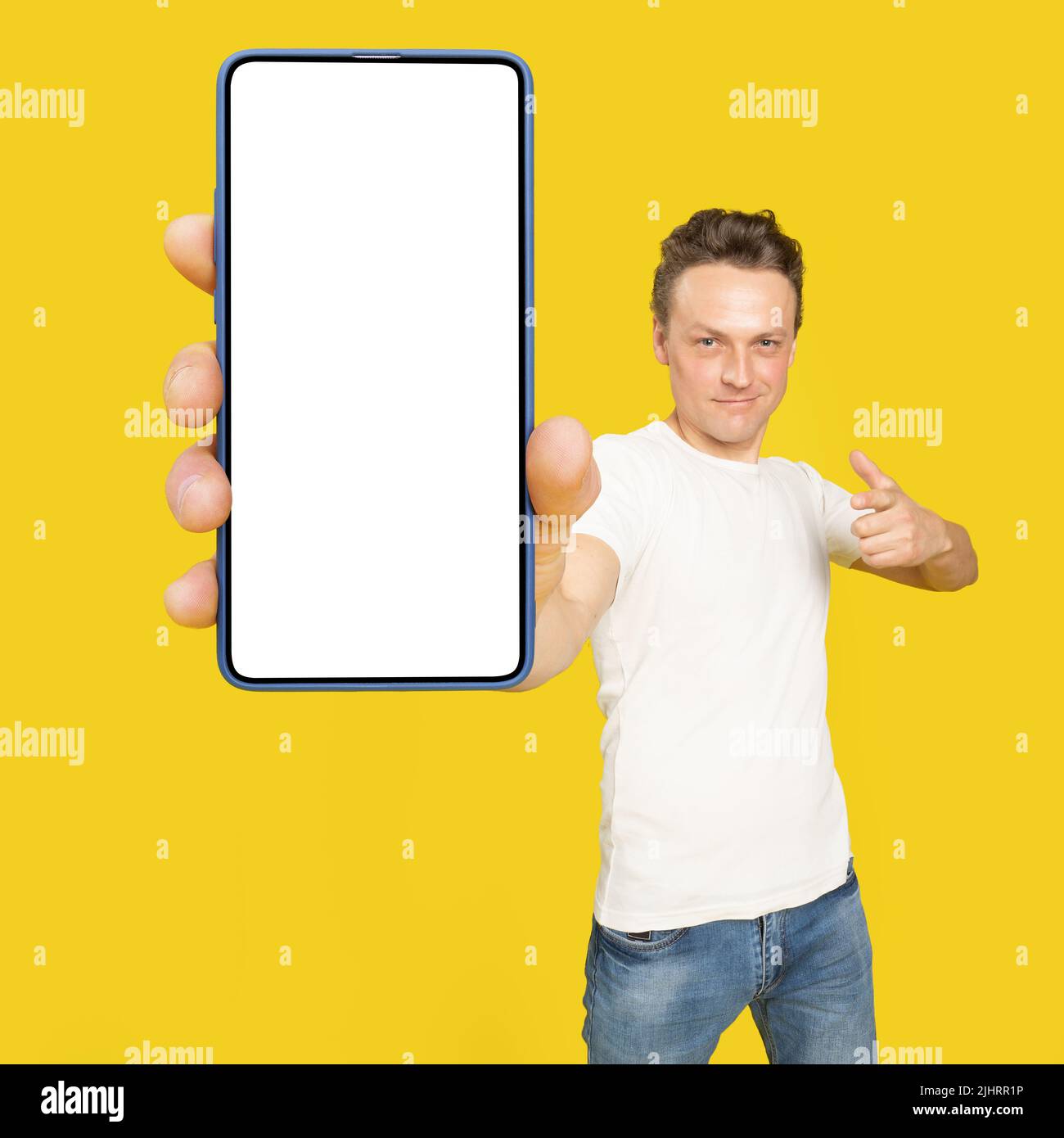Bell'uomo che tiene grande smartphone con schermo bianco che punta su uno schermo bianco vuoto, indossando maglietta bianca e jeans isolati su sfondo giallo. App per dispositivi mobili, grande offerta. Foto Stock