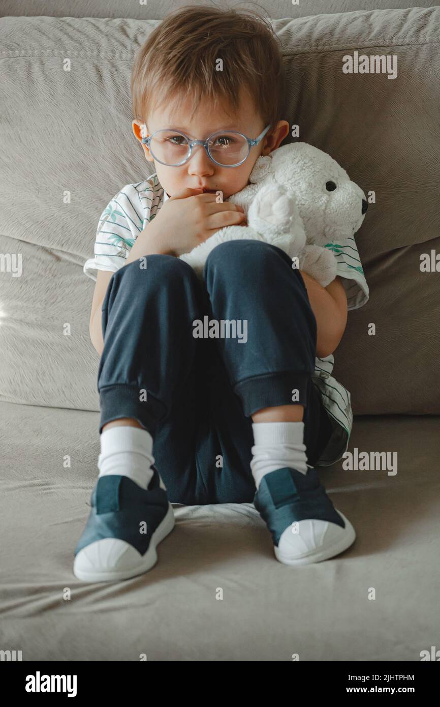 Un bambino con autismo in occhiali si siede sul divano e triste, abbraccia un s. Foto Stock