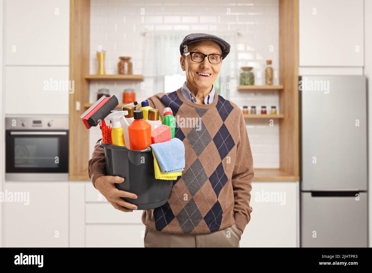 Uomo anziano che tiene un secchio con i rifornimenti di pulizia e sta in piedi all'interno di una cucina Foto Stock
