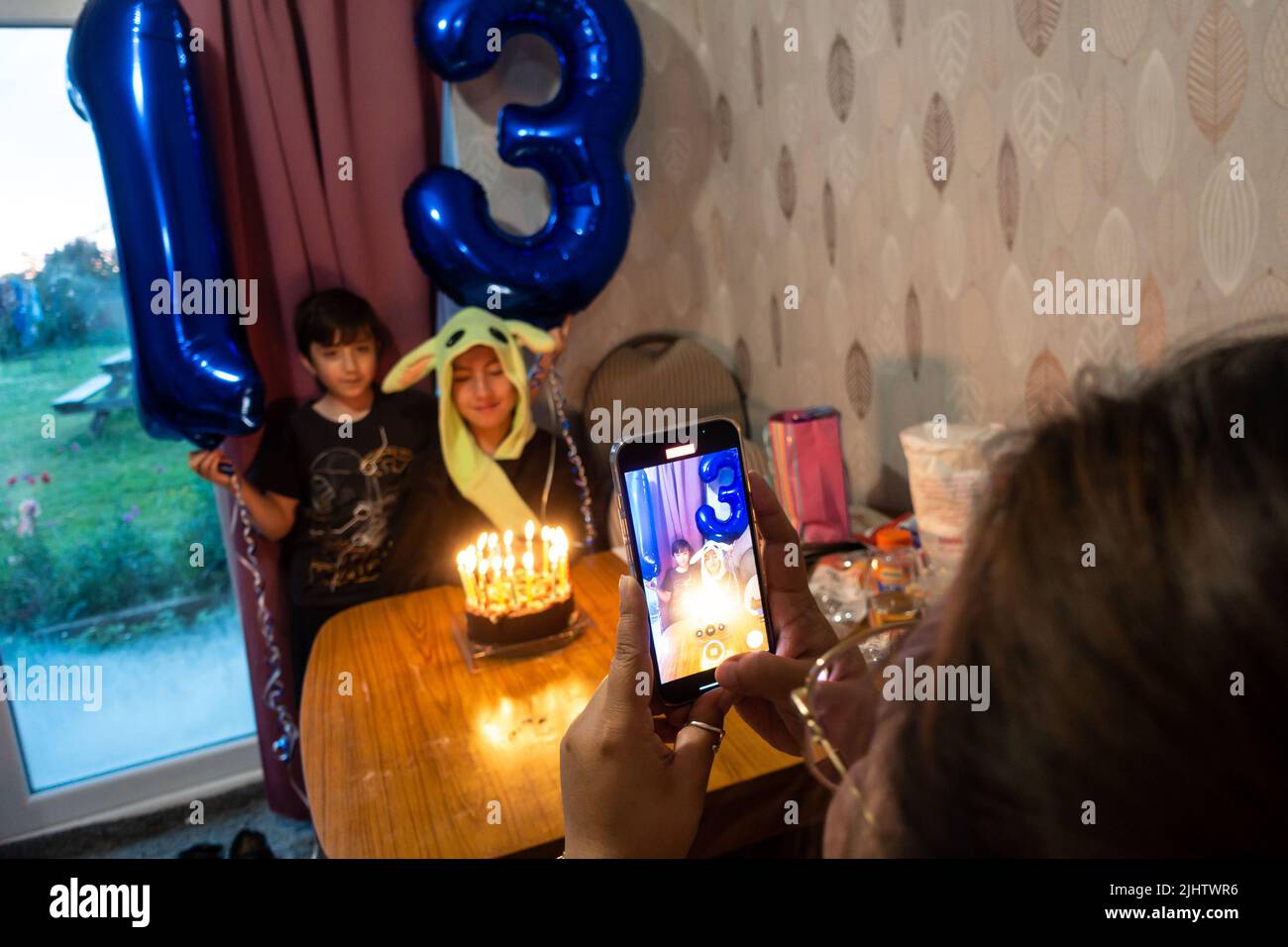 Una mamma scatta una foto con il cellulare dei suoi due figli che festeggiano il compleanno del figlio maggiore con palloncini di elio e torta di compleanno con candele. Foto Stock