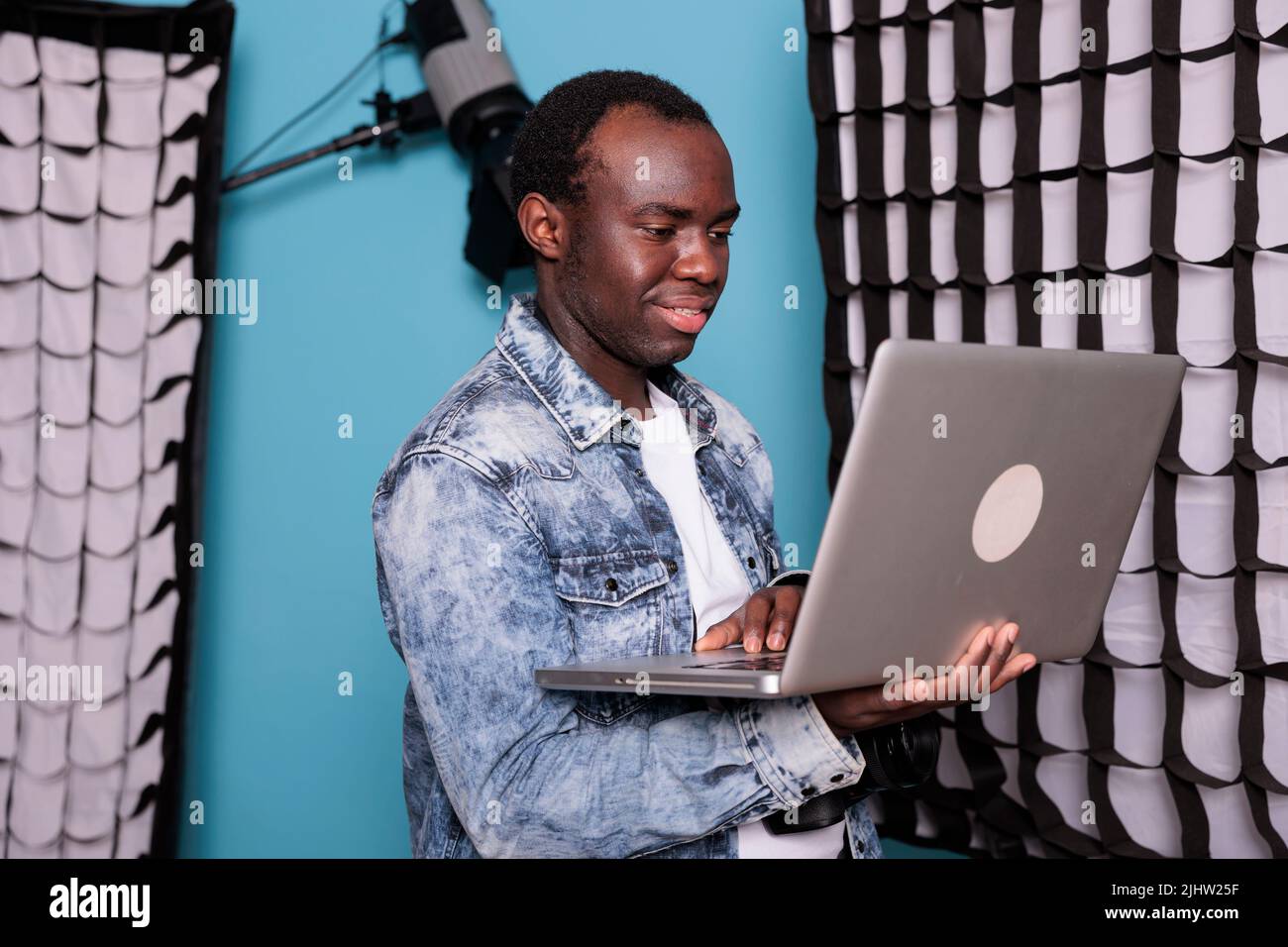 Gioioso direttore di produzione con fotocamera DSLR e computer portatile su sfondo blu. Fotografo sorridente che lavora su un computer portatile mentre si trova in uno studio dotato di luci e softbox professionali. Foto Stock