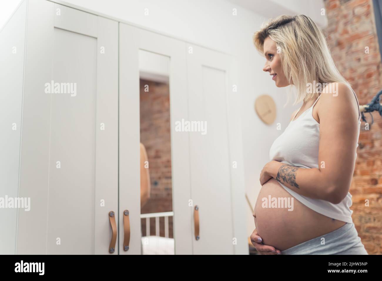 Buona gravidanza. La bionda caucasica millennial presto-essere madre in piedi davanti ad uno specchio del guardaroba e guardando il suo ventre grande di gravidanza. Foto di alta qualità Foto Stock