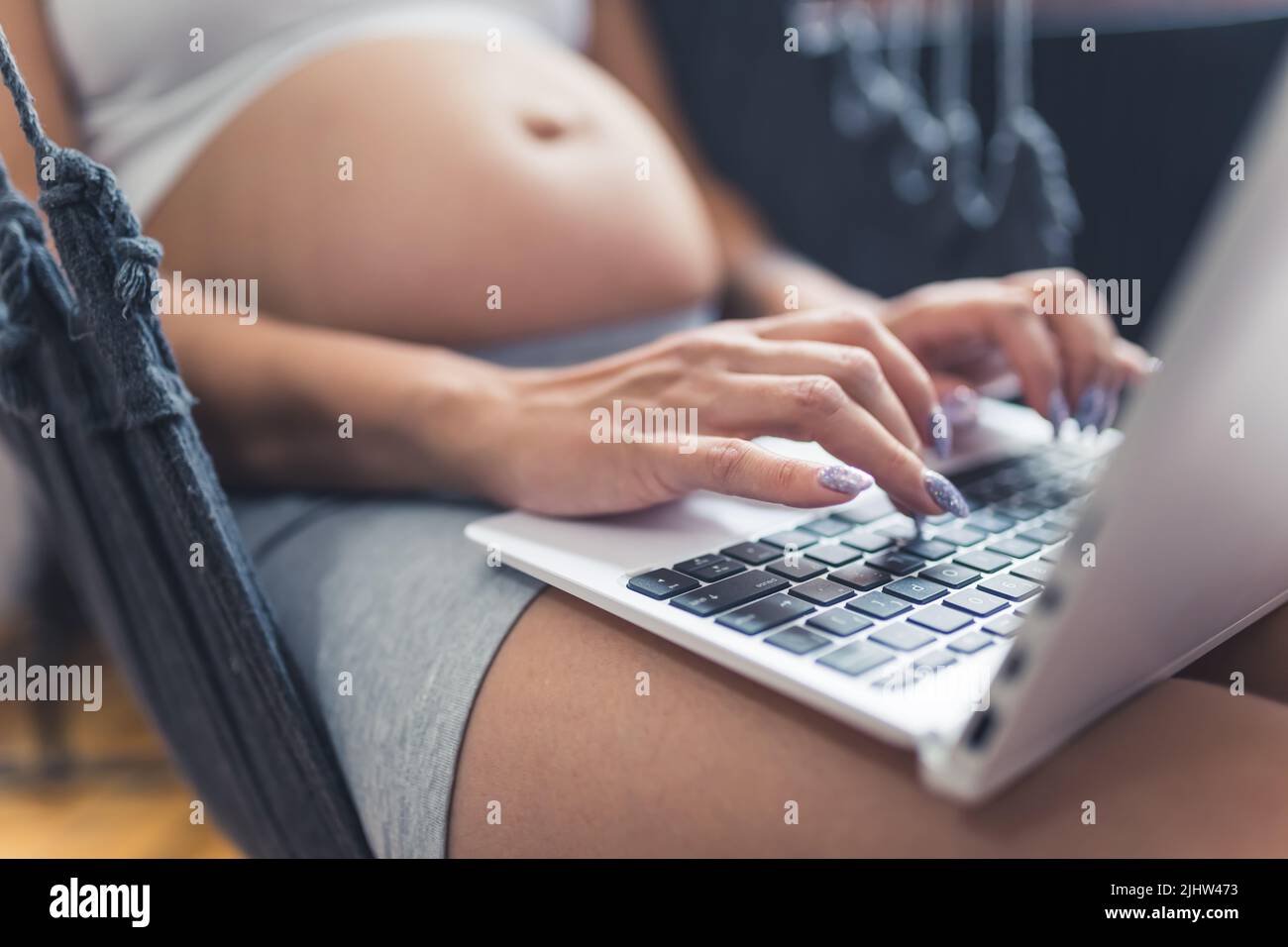 Persona incinta caucasica con lunghe unghie scintillanti che lavorano su un computer portatile mentre si siede in un'amaca interna. Foto di alta qualità Foto Stock