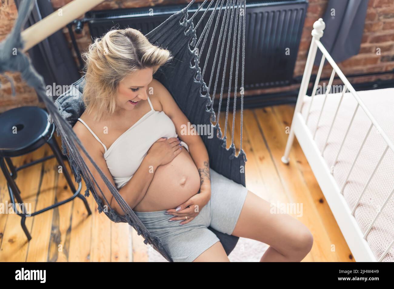 Girato dalla parte superiore di una giovane donna caucasica bionda con il ventre incinta che guarda il suo corpo, abbracciando il ventre, e sorridendo mentre si siede su un'amaca nera interna. Foto di alta qualità Foto Stock
