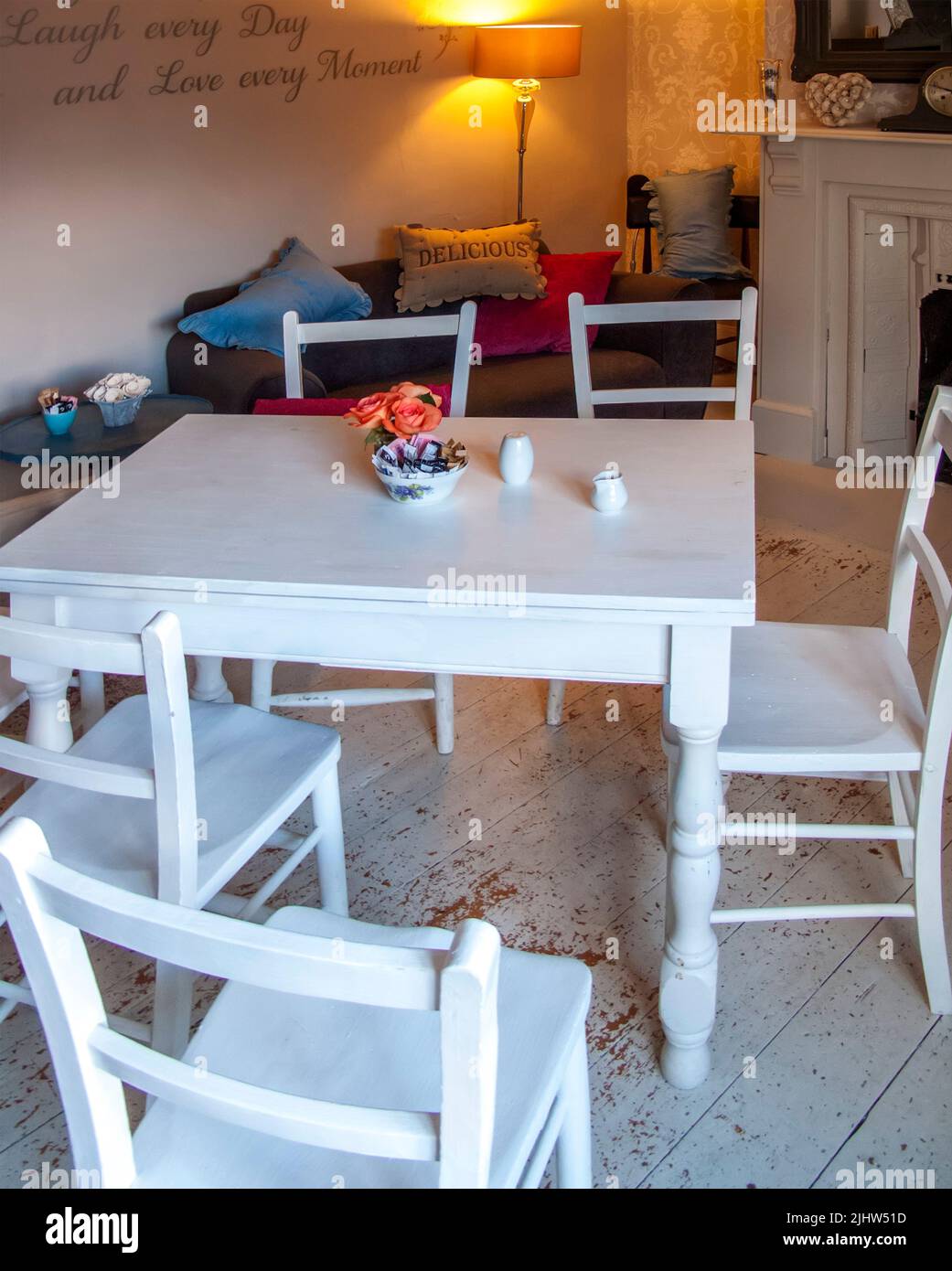 Camera con tavolo in legno bianco e sedie su pavimento in legno con lampada e camino e divano con cuscini Foto Stock