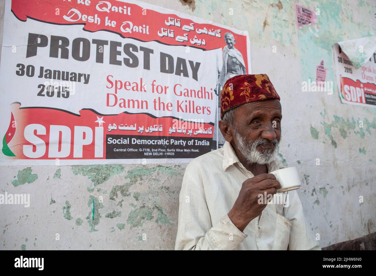 Un uomo (cieco in un occhio) beve una tazza di tè di fronte a un poster politico SDPI a Bijapur, India Foto Stock