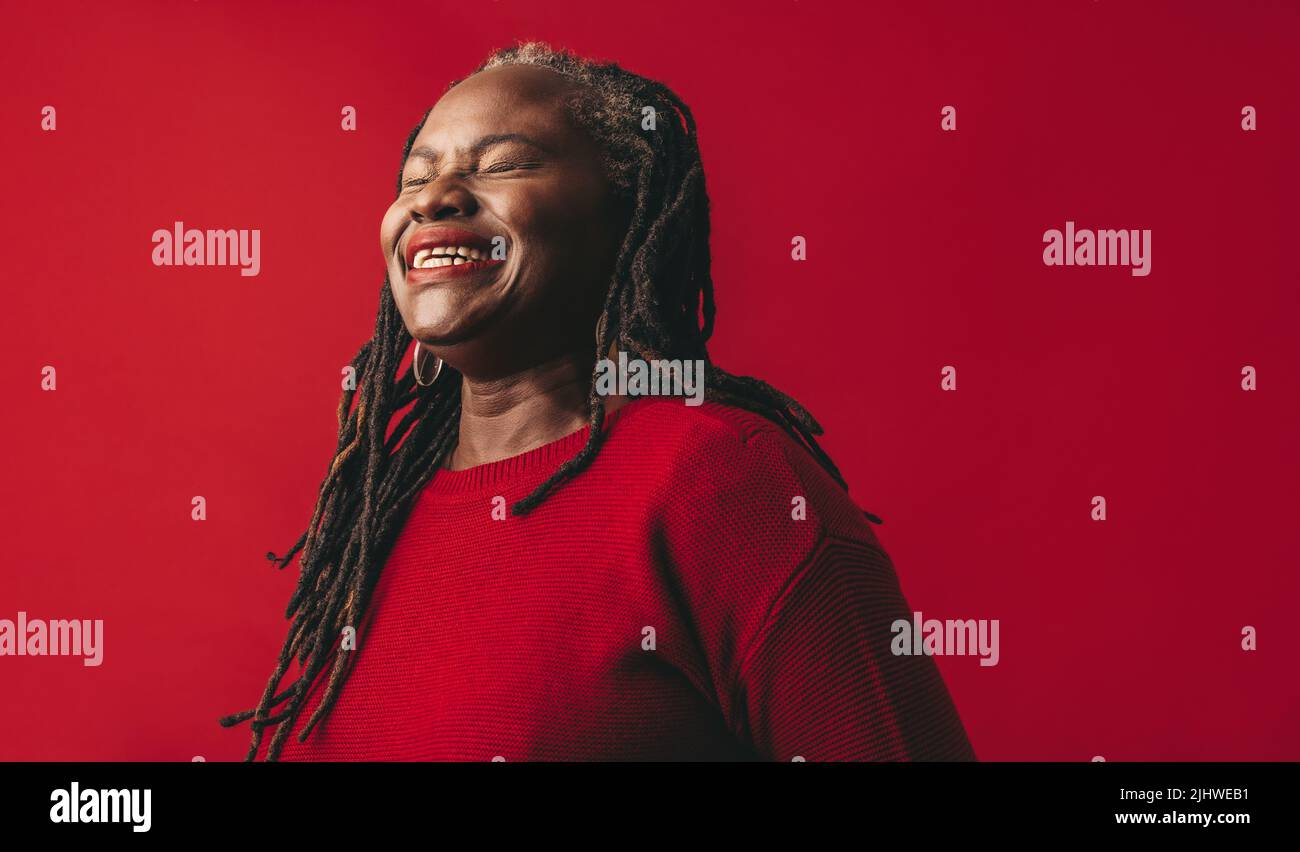 Donna elegante con dreadlocks sorridente gioiosamente mentre si alza su uno sfondo rosso. Donna matura felice abbracciando i suoi capelli naturali con orgoglio. Foto Stock