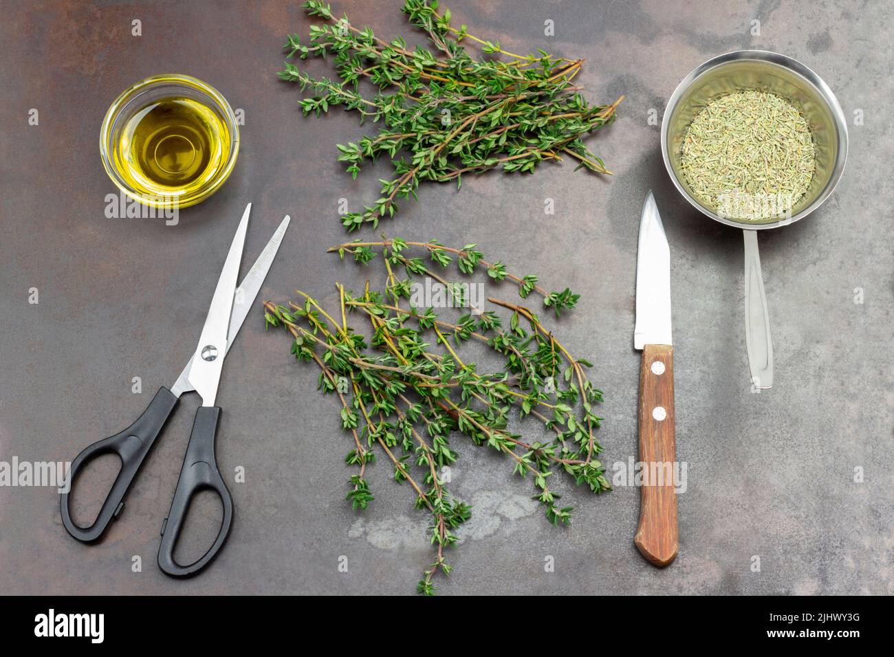 Forbici e coltello sul tavolo. Ciuffi di timo verde e timo secco in ciotola metallica. Olio nel recipiente di vetro. Top Vew. Fondo in metallo arrugginito. Foto Stock