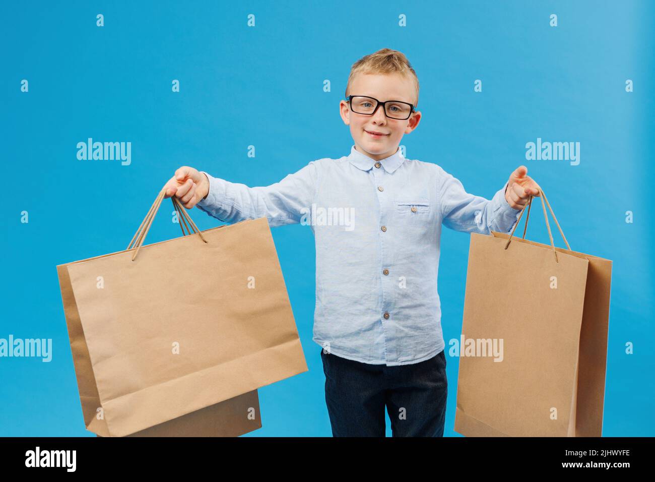 Simpatico ragazzo divertente in camicia blu che tiene buste di carta isolato su sfondo blu in studio. Concetto di stile di vita delle persone. Il ragazzo si offre di andare a fare shopping Foto Stock