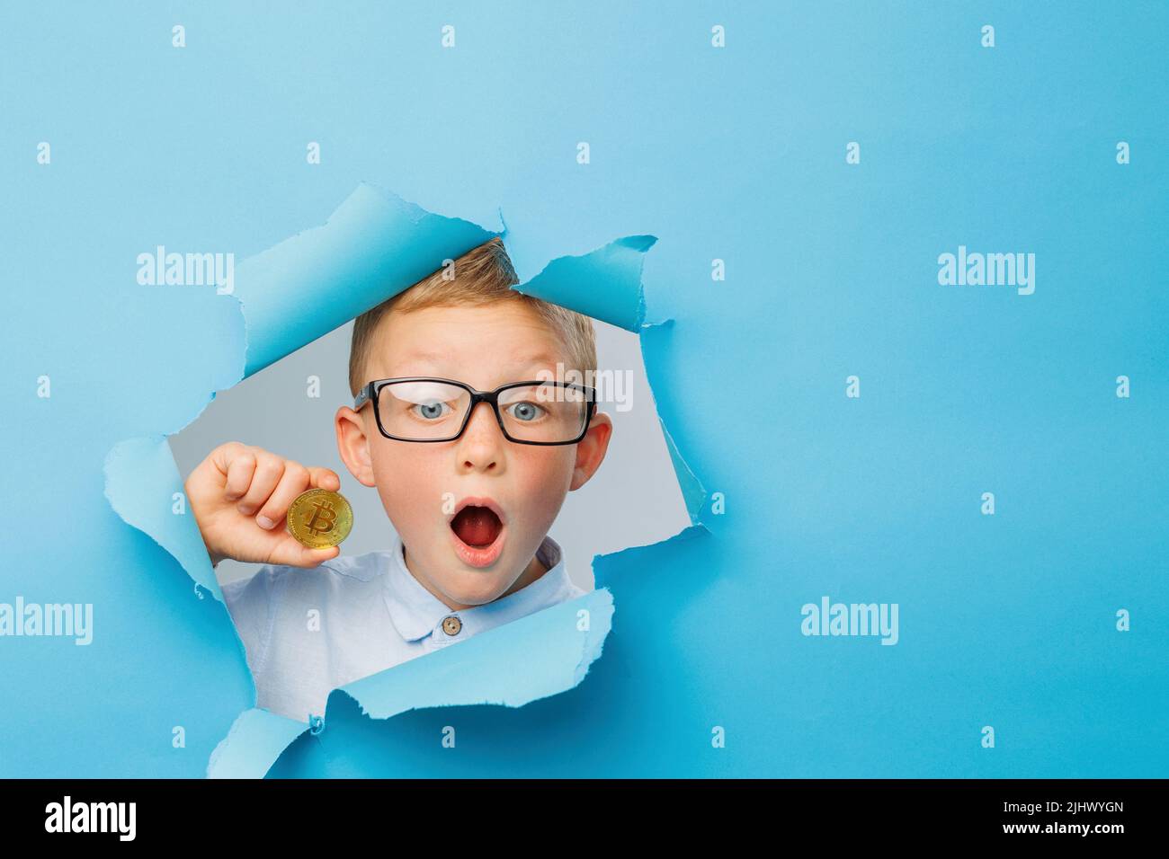 Felice simpatico businessboy in occhiali sta avendo divertimento e tenendo il bitcoin mano sul muro di fondo blu, si arrampica attraverso un foro nella carta. Luminoso e. Foto Stock