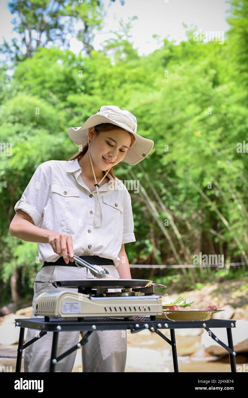 Attraente giovane donna asiatica arrostire una bistecca di manzo di maiale trita su una padella volante, cucinando con una stufa da picnic portatile. Barbecue all'aperto Foto Stock