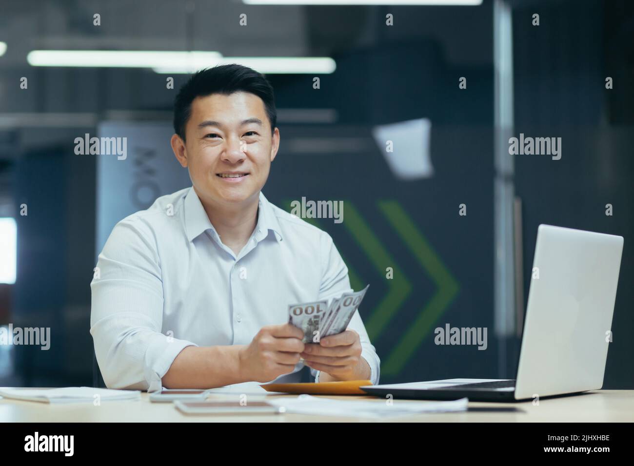 Ritratto di felice e riuscito uomo investitore mediatore asiatico sorridente e guardando la macchina fotografica che funziona in ufficio moderno che tiene soldi dei contanti dei dollari Foto Stock