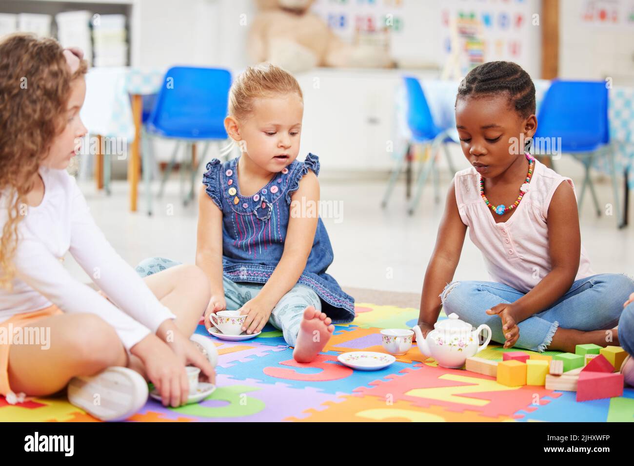 Gruppo di ragazze adorabili diverse seduti a gambe incrociate sul pavimento in preschool e avendo un tè partito. Tre bambini carini che giocano e si legano Foto Stock