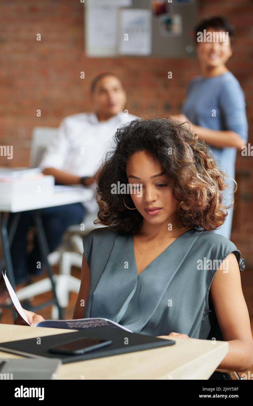 Leggendo l'ultima proposta di business, una giovane donna d'affari che passa attraverso la documentazione in un ufficio con i suoi colleghi in background. Foto Stock