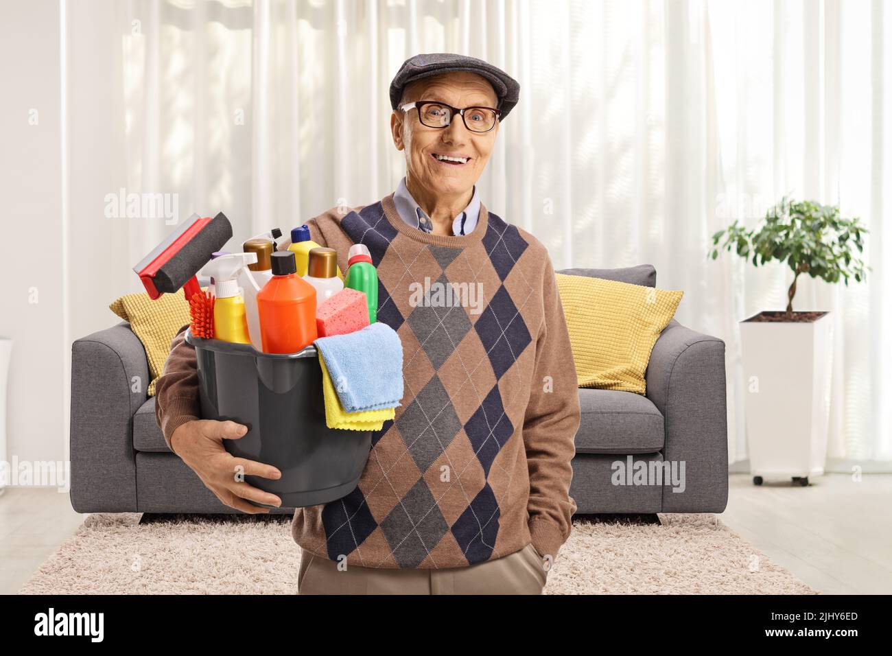 Uomo anziano che tiene un secchio con i rifornimenti di pulizia e sta in piedi all'interno di un soggiorno Foto Stock