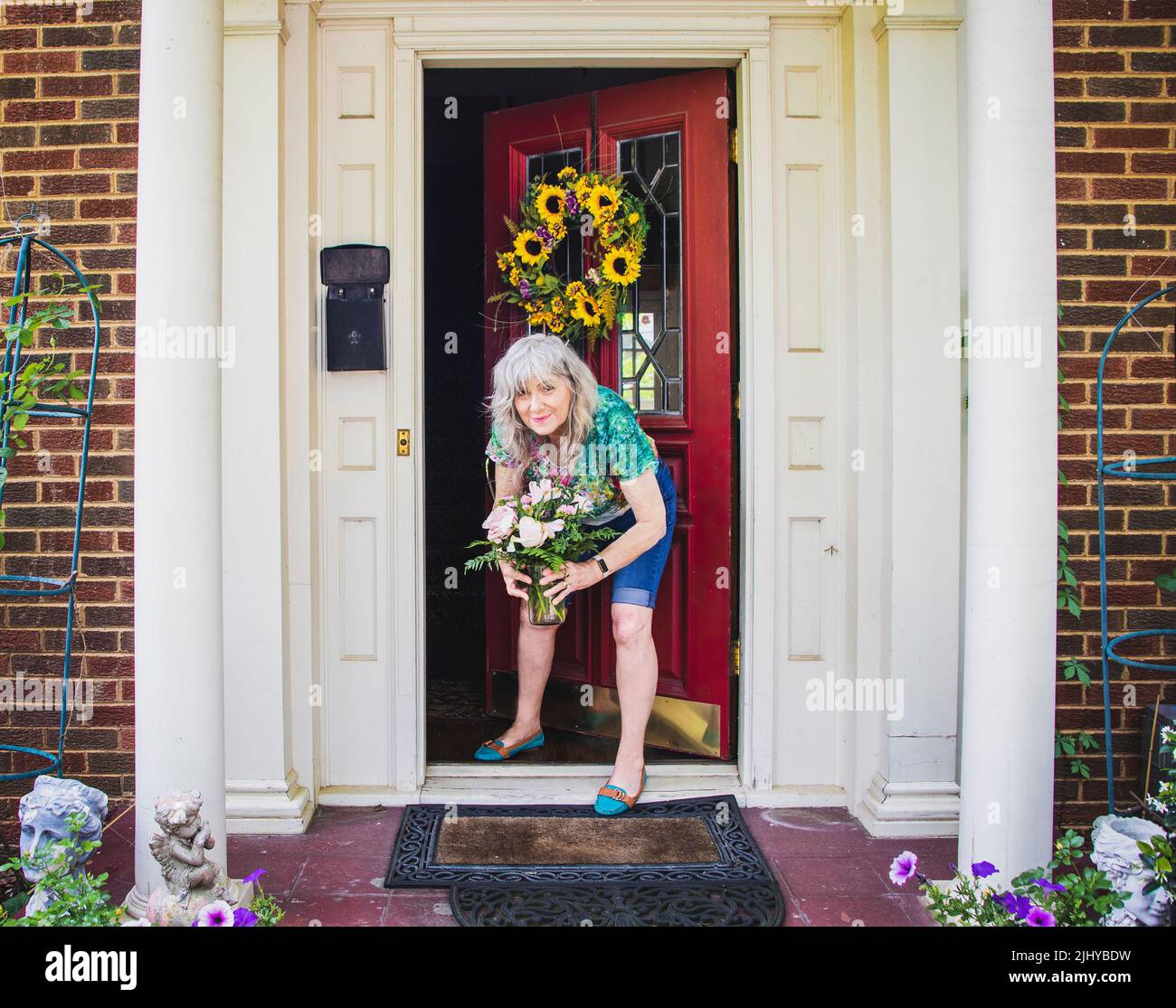 Donna anziana graziosa in shorts e t-shirt alla porta a casa mattone con colonne e corona di girasole raccoglie la consegna del fiore a sinistra sul portico per la nascita Foto Stock