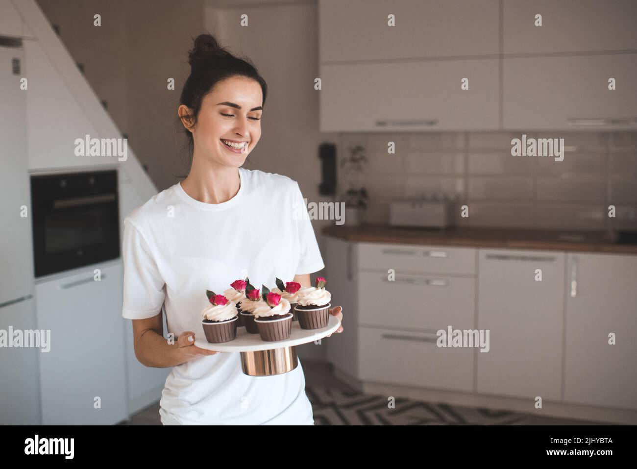 Sorridente bella donna felice facendo gustosi cupcake fatti in casa con fiore rosa decorazione vassoio di tenuta con pasticceria in cucina a casa. Foto Stock