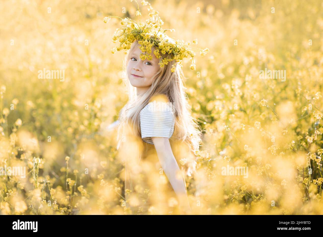 Carino ragazza elegante bambino 4-5 anni indossare wreath floreale stand in giallo prato verde all'aperto sulla natura sfondo sopra luce del sole. Infanzia. Estate Foto Stock