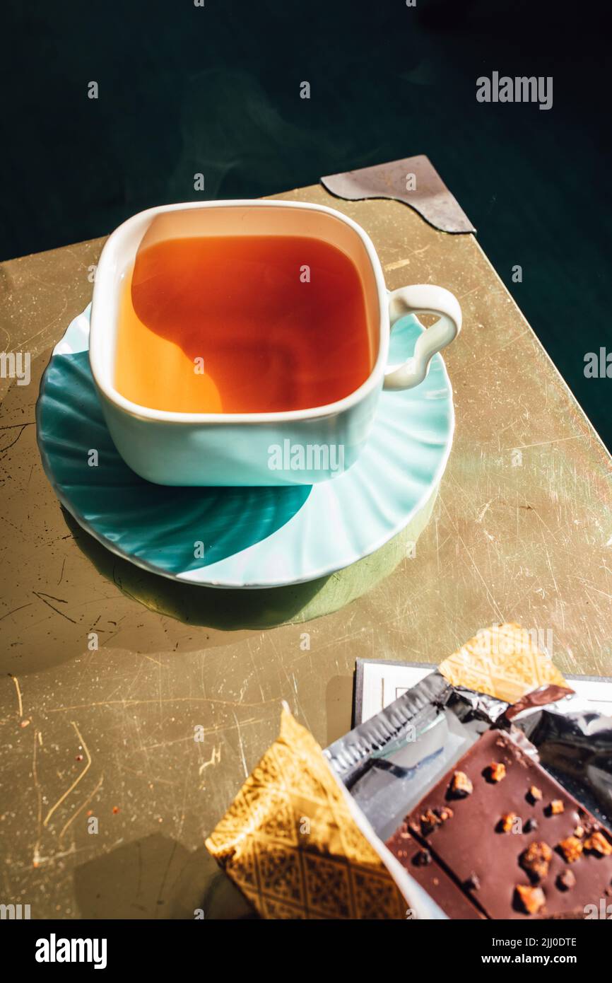 tè pomeridiano in teacup quadrato e piattino con cioccolato in involucro d'oro Foto Stock
