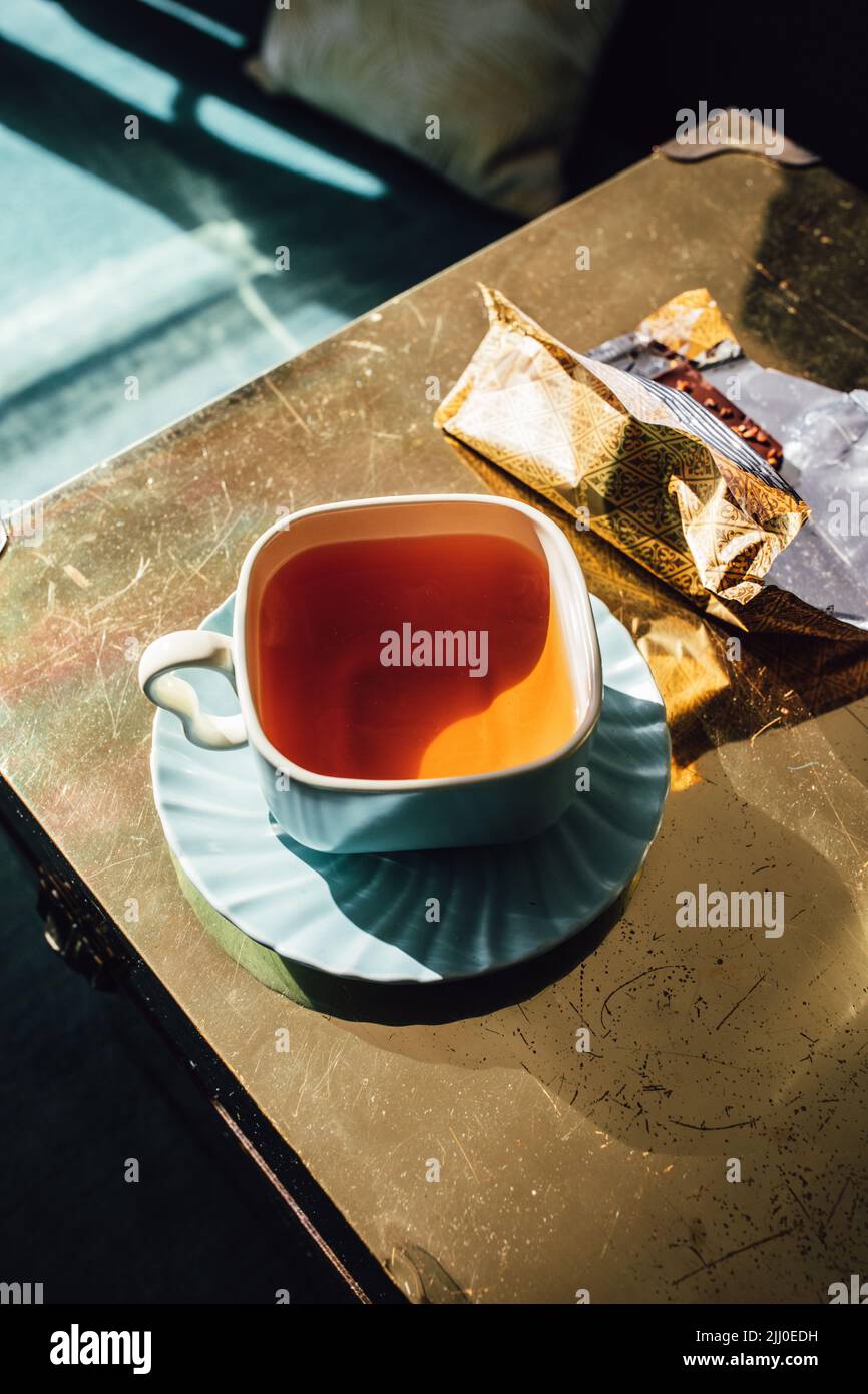 tè pomeridiano in teacup quadrato e piattino con cioccolato in involucro d'oro Foto Stock