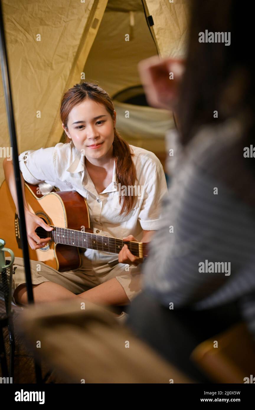 Attraente giovane asiatico femmina godere di suonare la chitarra e cantare con i suoi amici, avendo una festa falò di notte nel campeggio. Foto Stock
